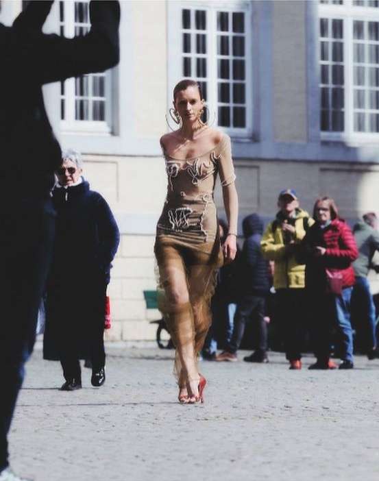 A slender woman with large hoop earrings walks in a sheer dress and high heels on a cobblestone surface, surrounded by onlookers.