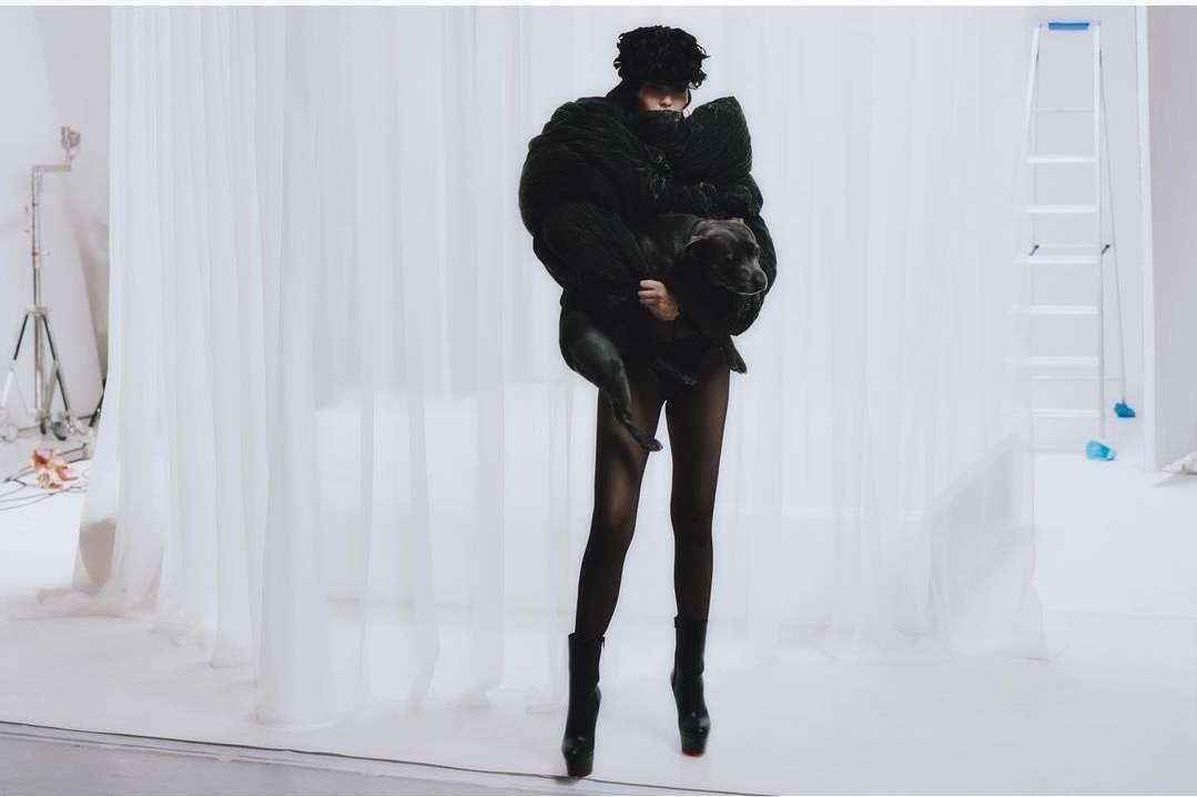 Woman in black coat holds a dog in a photo studio with white backdrop and lighting equipment, creating a striking fashion image.