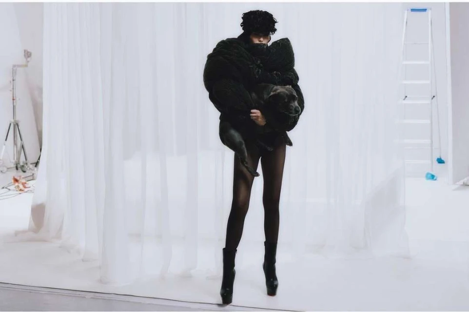 Woman in black coat holds a dog in a photo studio with white backdrop and lighting equipment, creating a striking fashion image.