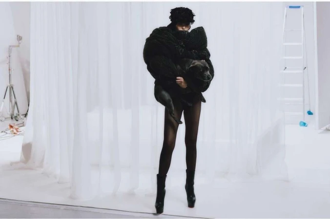 Woman in black coat holds a dog in a photo studio with white backdrop and lighting equipment, creating a striking fashion image.