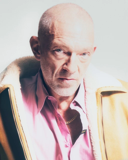 Close-up portrait of a fair-skinned adult male with a shaved head, wearing a pink shirt and a tan shearling-lined jacket against a white backdrop.