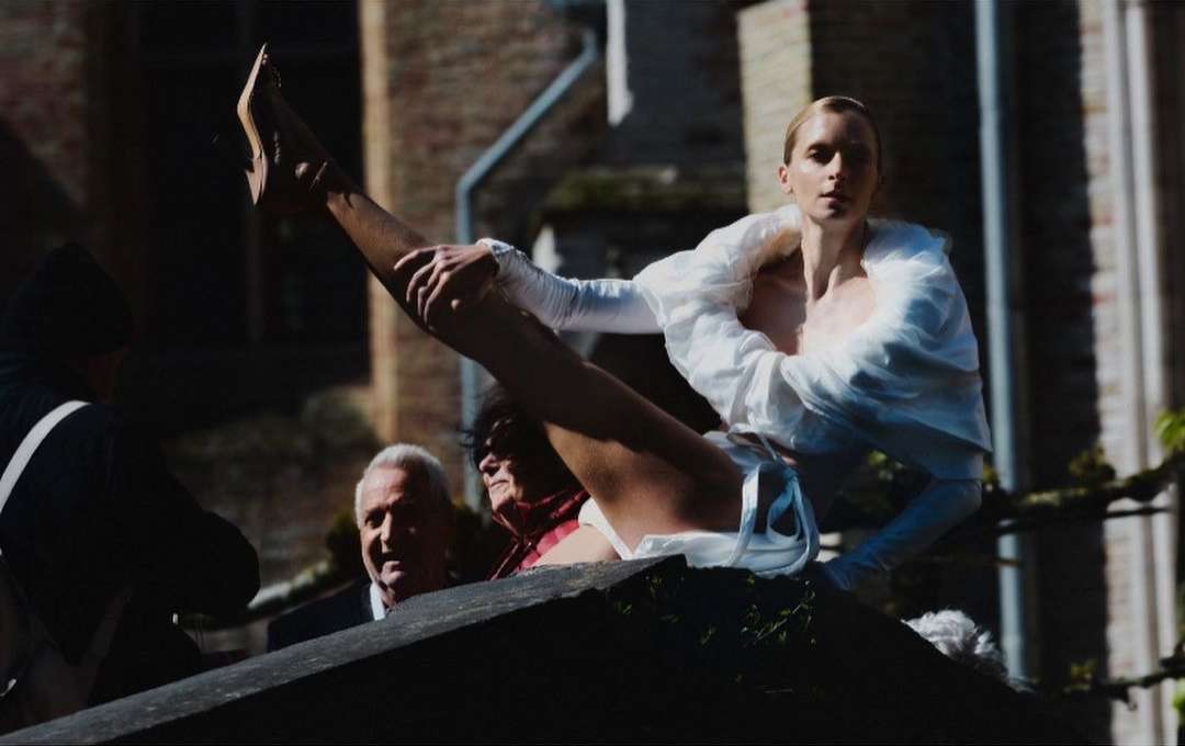 Woman in white outfit does leg extension on ledge with bystanders. Image with diffused lighting, high angle on stone building.