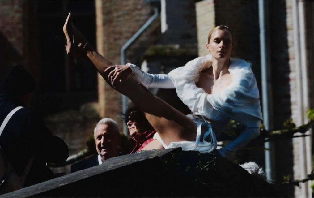 Woman in white outfit does leg extension on ledge with bystanders. Image with diffused lighting, high angle on stone building.