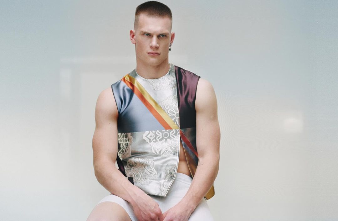 A young male model with a buzz cut wearing a sleeveless patterned top against a white seamless background