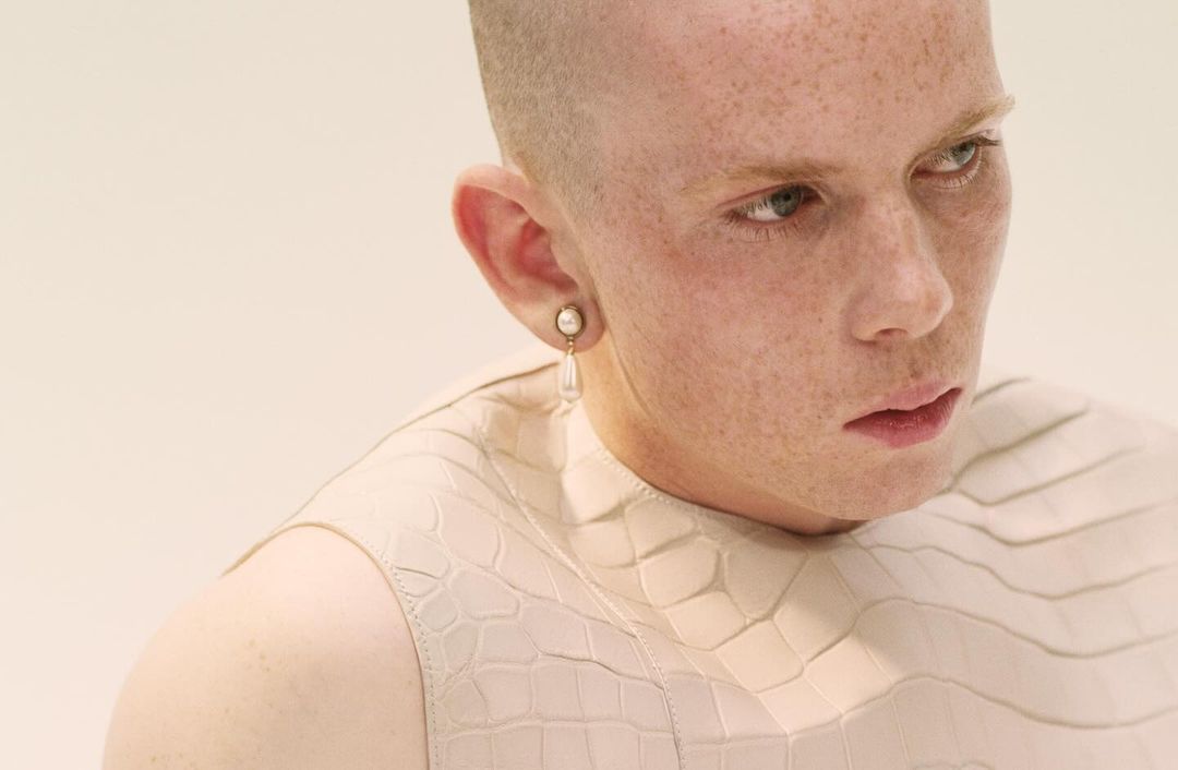Close-up portrait of a fair-skinned adult with a shaved head wearing a textured light-beige top and pearl earring against an off-white background.