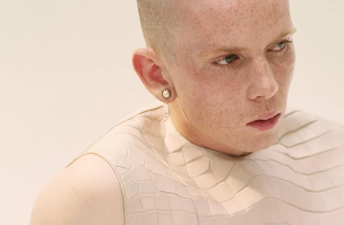Close-up portrait of a fair-skinned adult with a shaved head wearing a textured light-beige top and pearl earring against an off-white background.