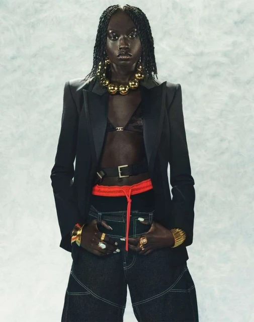 Fashion portrait of a Black woman wearing a black blazer, black top, and jeans, accessorized with gold jewelry, against a mottled backdrop.