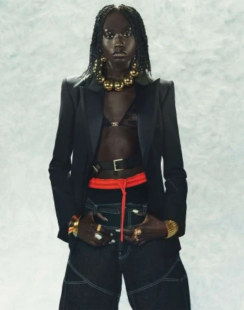 Fashion portrait of a Black woman wearing a black blazer, black top, and jeans, accessorized with gold jewelry, against a mottled backdrop.