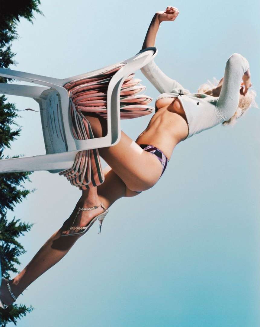 Fashion shot of a woman in a bikini and jacket posing with a plastic chair against a blue sky with trees partially visible.