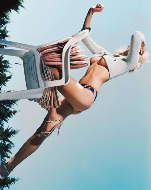 Fashion shot of a woman in a bikini and jacket posing with a plastic chair against a blue sky with trees partially visible.
