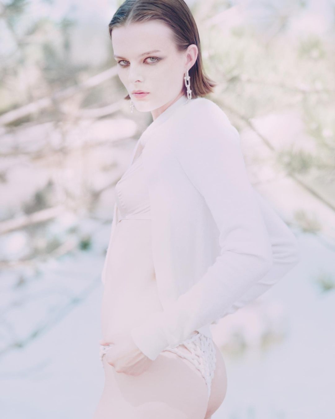 Adult female poses in a white long-sleeved shirt and underwear against a blurry winter forest backdrop, styled with silver earrings.