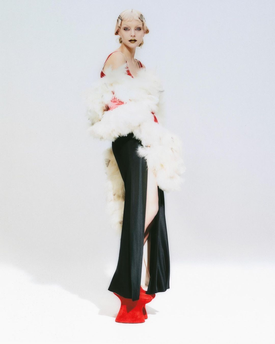 Fashionable woman in black dress, feather boa, and red shoes against white backdrop in studio fashion shot, editorial composition.