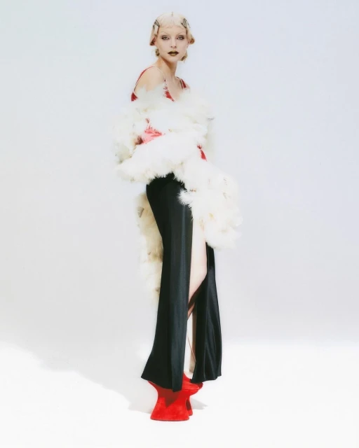 Fashionable woman in black dress, feather boa, and red shoes against white backdrop in studio fashion shot, editorial composition.