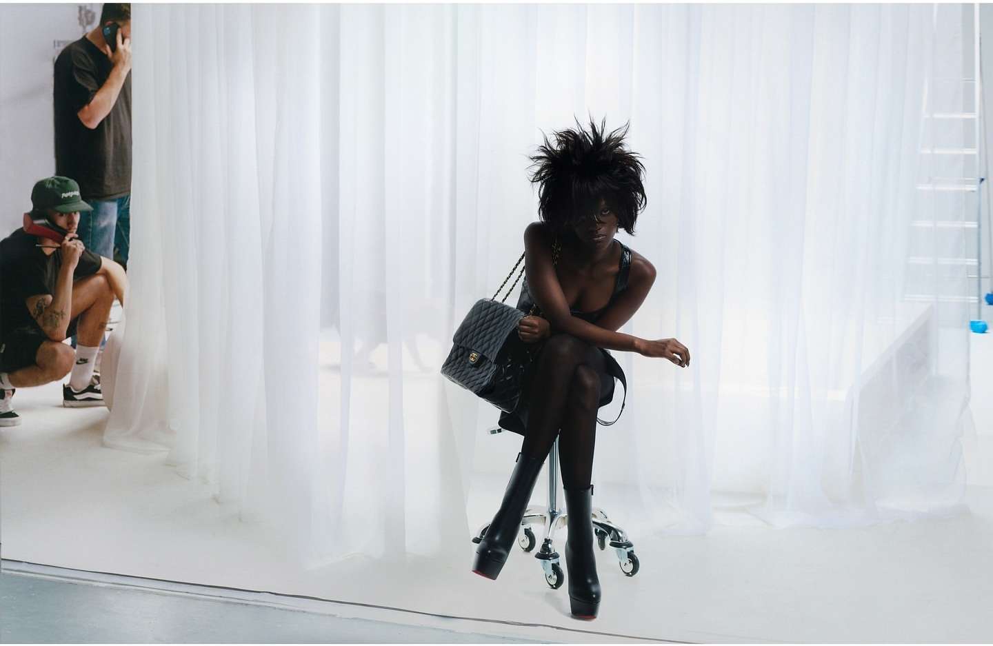 An adult female poses in a photo studio, seated on a rolling chair with a quilted leather bag, wearing platform boots and a black dress.