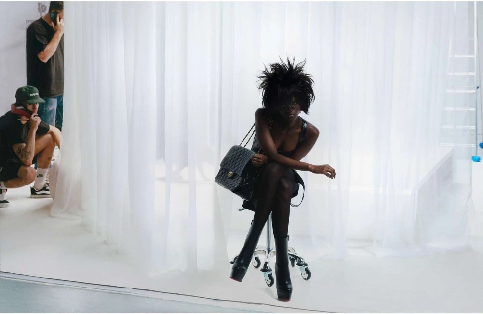 An adult female poses in a photo studio, seated on a rolling chair with a quilted leather bag, wearing platform boots and a black dress.