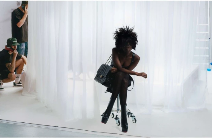 An adult female poses in a photo studio, seated on a rolling chair with a quilted leather bag, wearing platform boots and a black dress.