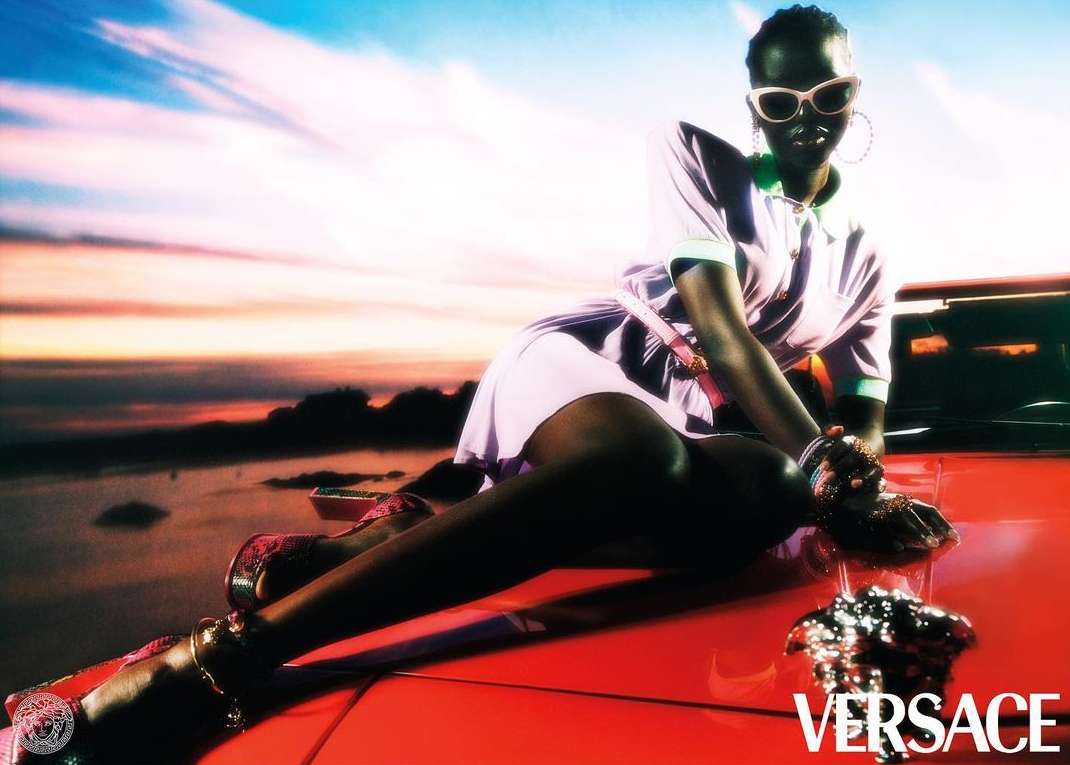 A dark-skinned fashion model poses on the hood of a red car in a light purple dress with sunglasses, bracelets, and hoop earrings.