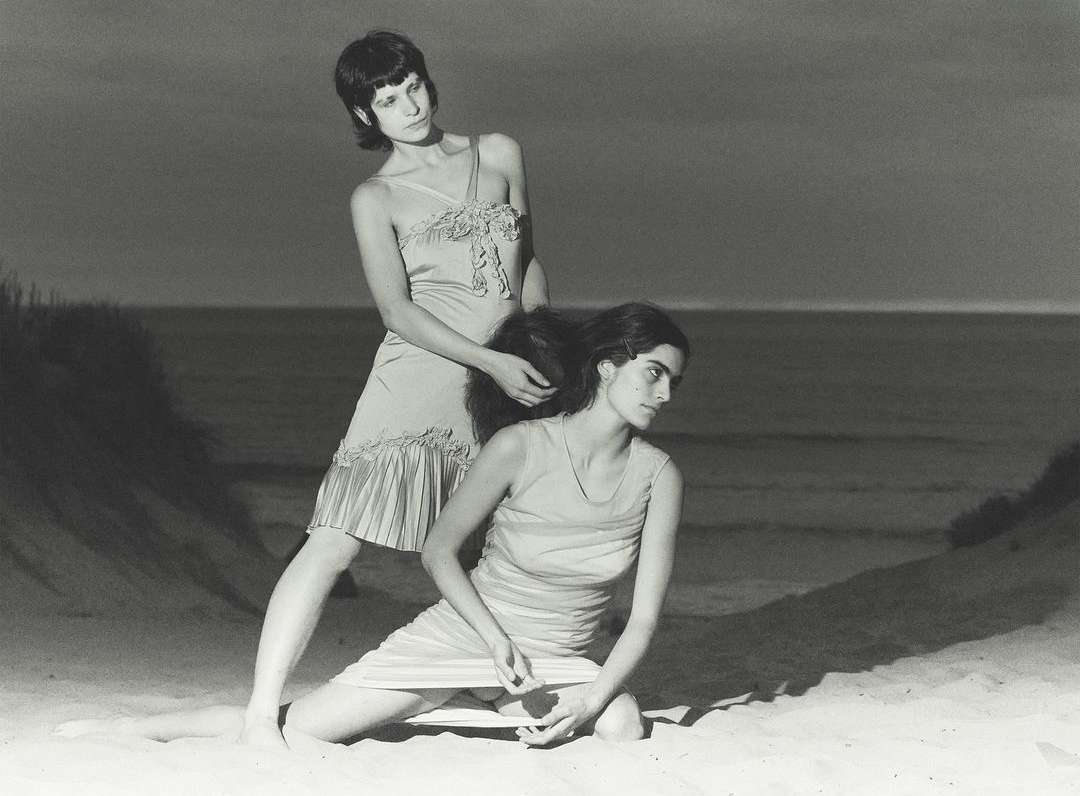 Two adult females pose on a beach with ocean background. One woman styles the other's hair in a vintage, black-and-white outdoor portrait.