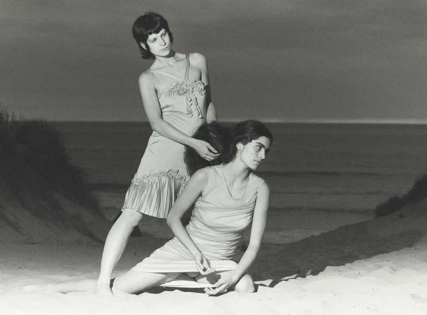 Two adult females pose on a beach with ocean background. One woman styles the other's hair in a vintage, black-and-white outdoor portrait.