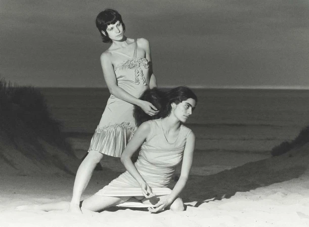 Two adult females pose on a beach with ocean background. One woman styles the other's hair in a vintage, black-and-white outdoor portrait.