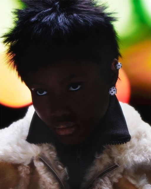 Close-up portrait of an adult with dark skin, wearing a light-colored furry jacket and studded earrings, against a blurred colorful background.