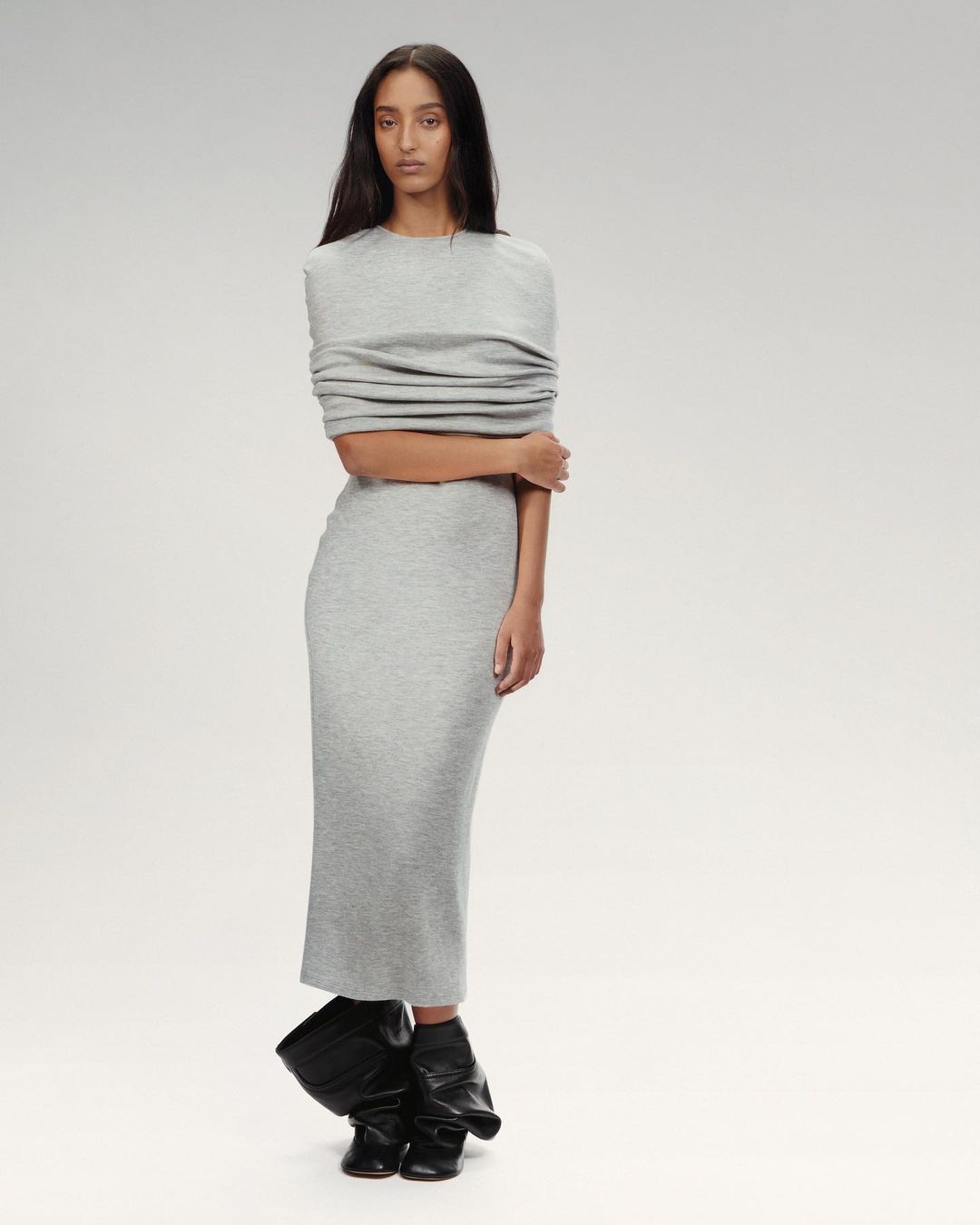 A model on a runway wears a grey midi-length dress with draped shoulder detail and black statement boots against a neutral background.