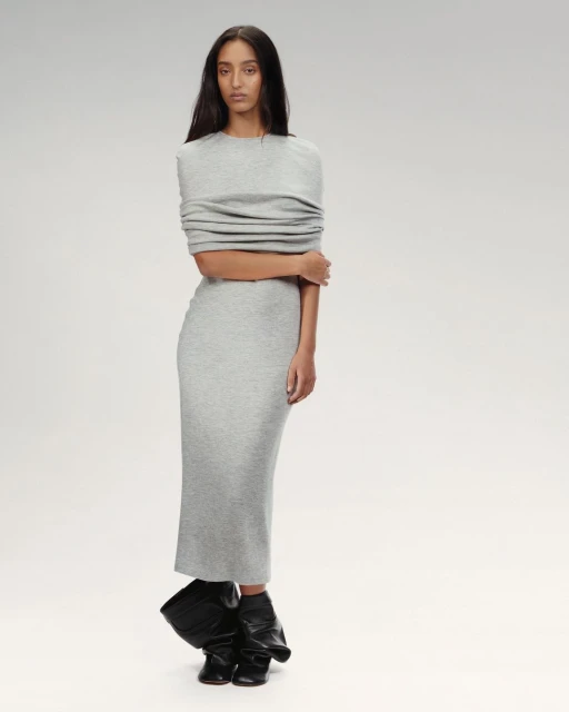 A model on a runway wears a grey midi-length dress with draped shoulder detail and black statement boots against a neutral background.