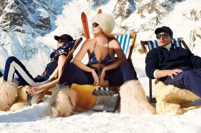Three people in swimwear and winter apparel lounge in beach chairs on a snowy mountainside with skis; winter fashion editorial shoot.