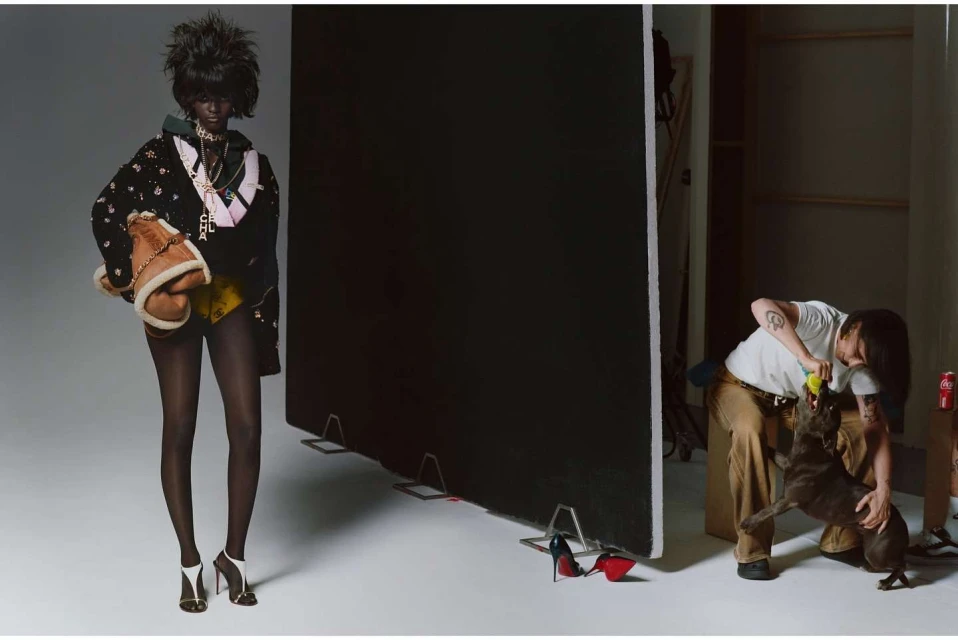 Fashion model in a black jacket and yellow shorts poses in a studio, while a photographer plays with a dog in the background.