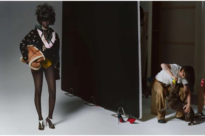 Fashion model in a black jacket and yellow shorts poses in a studio, while a photographer plays with a dog in the background.