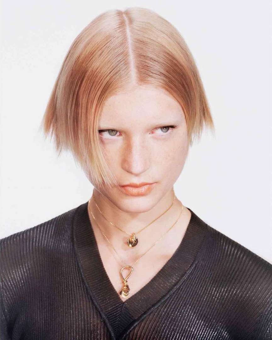 Portrait of a blonde woman wearing a black ribbed v-neck top and gold pendant necklaces against a white background.