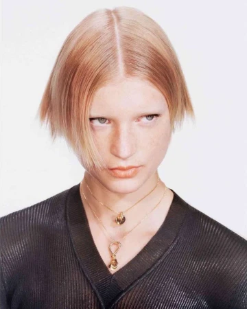 Portrait of a blonde woman wearing a black ribbed v-neck top and gold pendant necklaces against a white background.