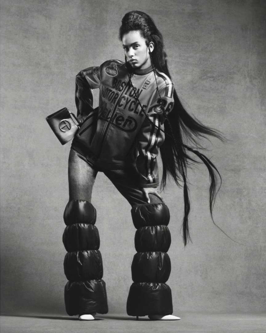 Monochrome fashion shot of a young adult woman with long hair wearing puffy pants and a motorcycle racing jacket in a studio.