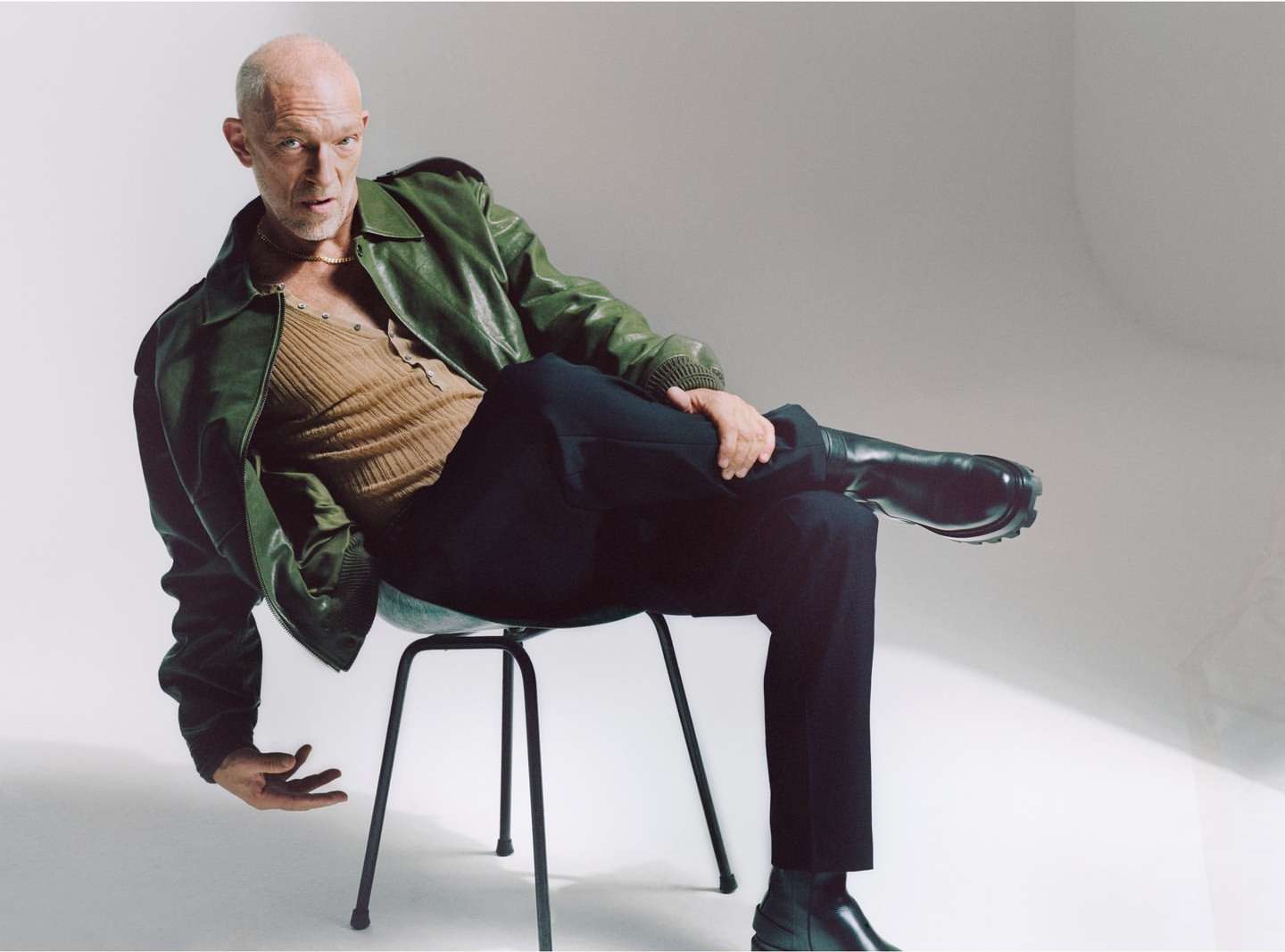 An adult male sits on a black stool in a photo studio, wearing a green leather jacket, brown shirt, black pants, and black boots.