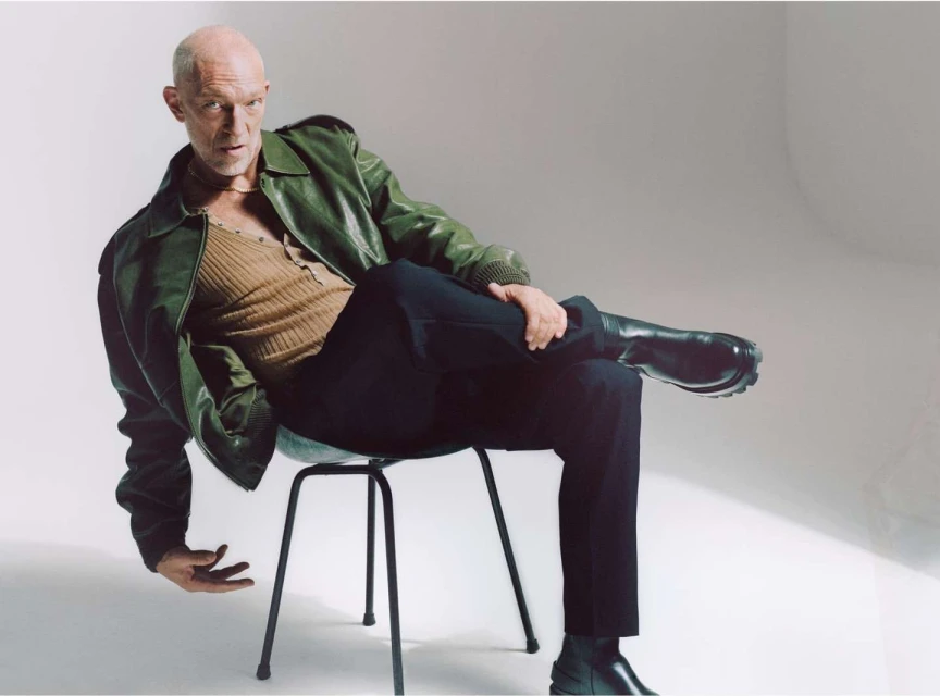 An adult male sits on a black stool in a photo studio, wearing a green leather jacket, brown shirt, black pants, and black boots.