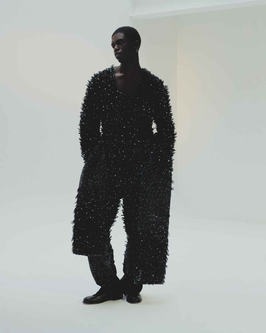 Black man wearing a heavily textured dark fringed outfit standing against a plain white wall