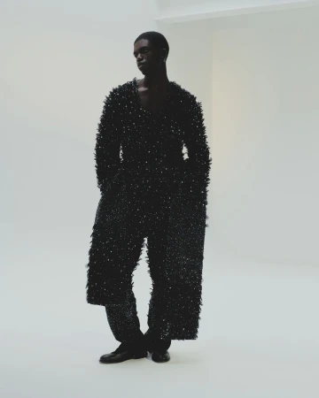 Black man wearing a heavily textured dark fringed outfit standing against a plain white wall