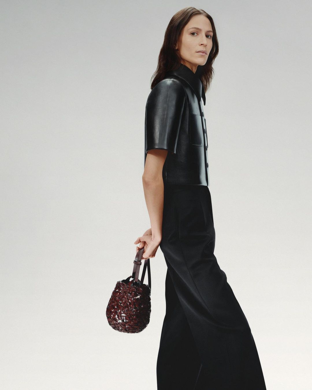 Fashionable adult female model wearing a black leather shirt and wide leg pants, holding a beaded dark red handbag in a studio setting.