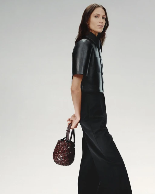 Fashionable adult female model wearing a black leather shirt and wide leg pants, holding a beaded dark red handbag in a studio setting.