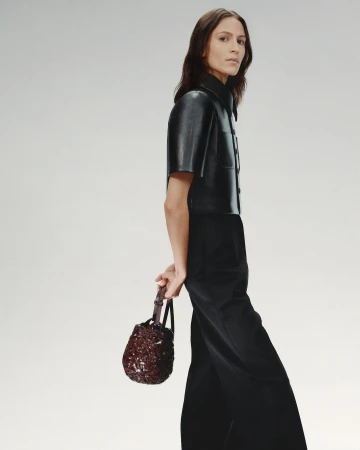 Fashionable adult female model wearing a black leather shirt and wide leg pants, holding a beaded dark red handbag in a studio setting.