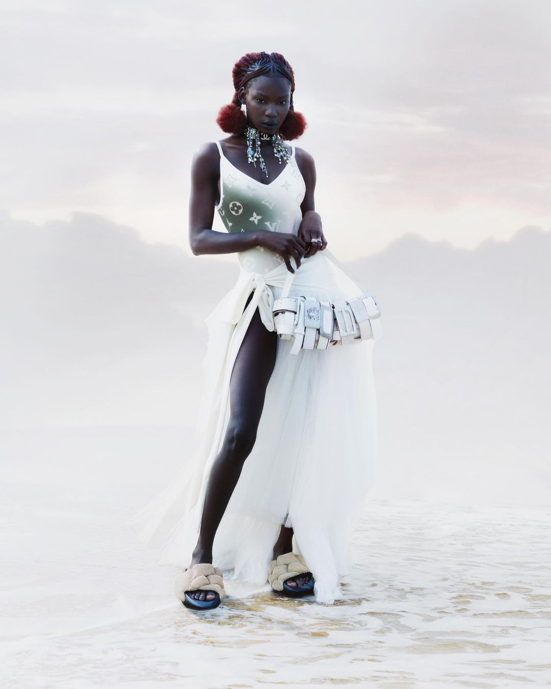 An African female model with braided hair and earrings is posing in the shallow water wearing a dress on a cloudy day near the sea.