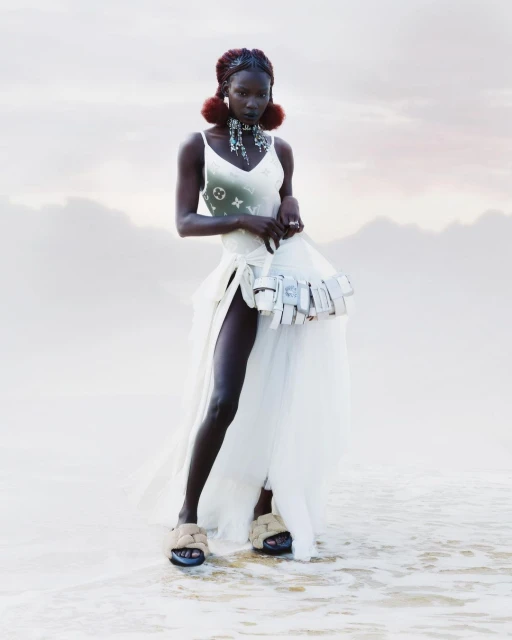 An African female model with braided hair and earrings is posing in the shallow water wearing a dress on a cloudy day near the sea.