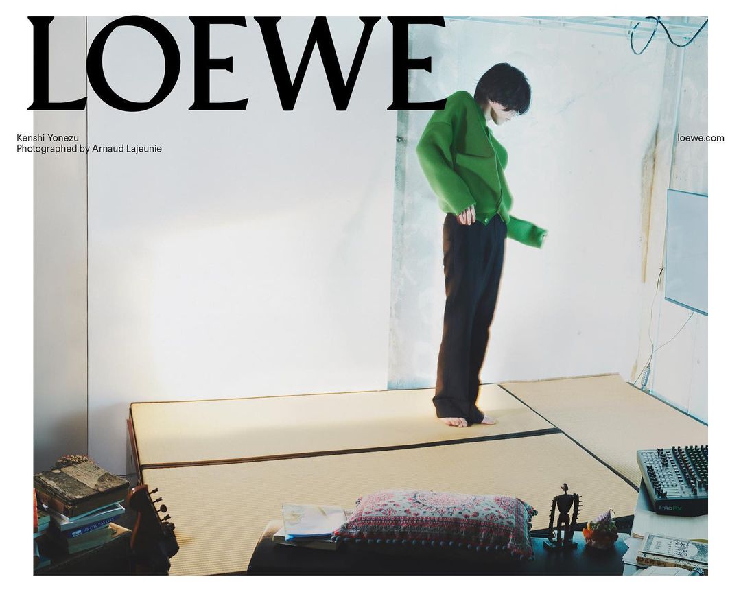 An Asian person with dark hair poses in a lime green sweater and dark trousers on a platform in a minimalist studio setting with soft lighting.
