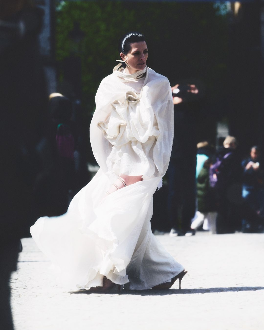 Fashion model in a voluminous white dress walks on a paved surface, surrounded by blurred onlookers, capturing a moment of runway elegance.