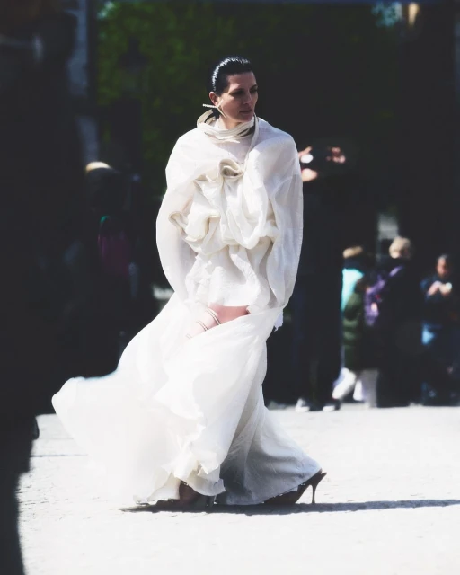 Fashion model in a voluminous white dress walks on a paved surface, surrounded by blurred onlookers, capturing a moment of runway elegance.