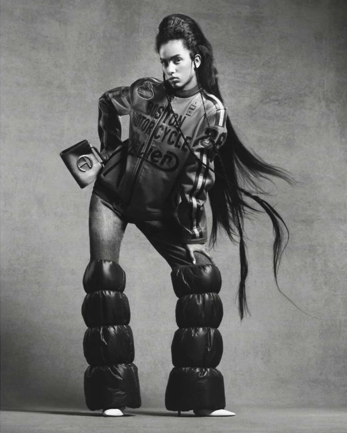 Model with long hair wearing a printed leather jacket and padded boots holding a handbag against a textured backdrop