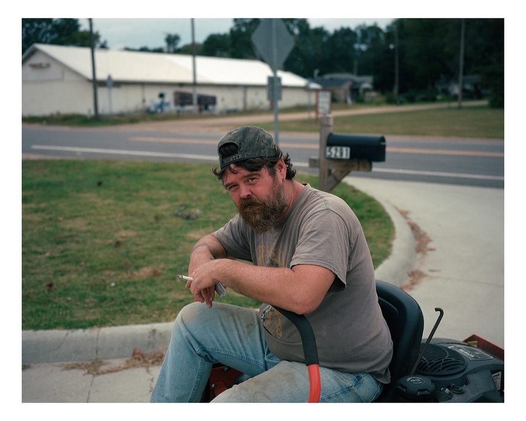 A man with a beard in a gray t-shirt and baseball cap sits on a lawn mower, smoking a cigarette on a suburban street in the daytime.