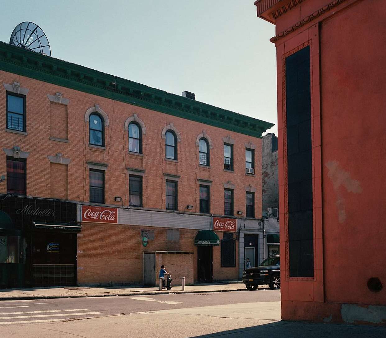 An urban street scene features brick buildings, Coca-Cola signage, and a man on a scooter on a sunny day, providing a glimpse into city life.