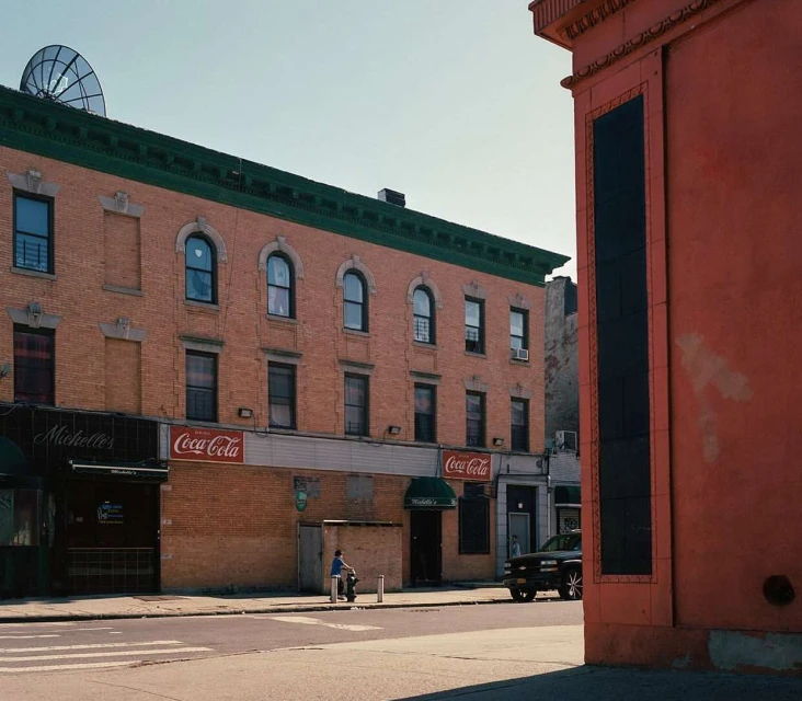 An urban street scene features brick buildings, Coca-Cola signage, and a man on a scooter on a sunny day, providing a glimpse into city life.
