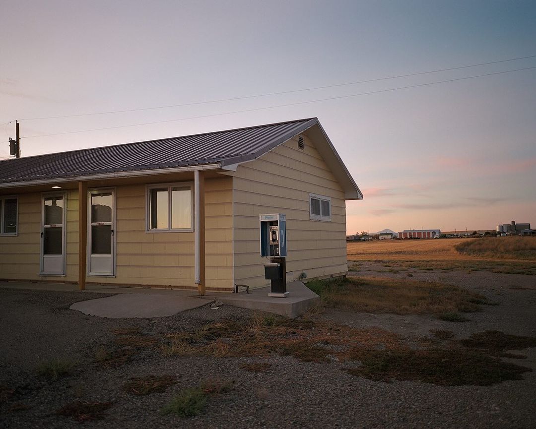 Exterior view of a roadside motel featuring a vintage payphone booth on a gravel lot in a rural landscape during a warm sunset.